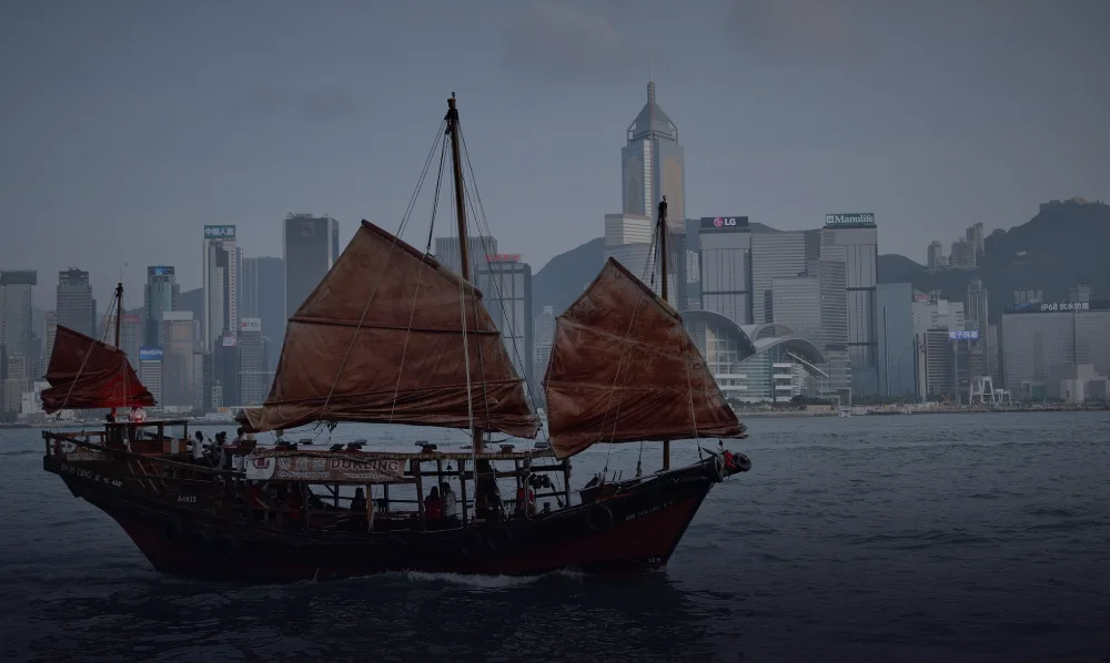 Colour photograph of the junk Dukling under rust-red sails, the Central skyline behind