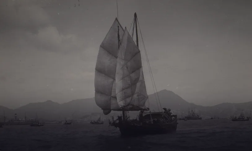 Black-and-white photograph of a wooden junk under full sail in the harbour, hills and steamships behind